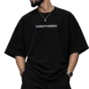 Male fashion model wearing an oversized black T-shirt with “UNBOTHERED” printed in white, styled with black cargo pants, sunglasses, a silver chain, watch, and ear piercing in a dark studio setting.