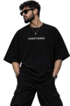 Male fashion model wearing an oversized black T-shirt with “UNBOTHERED” printed in white, styled with black cargo pants, sunglasses, a silver chain, watch, and ear piercing in a dark studio setting.