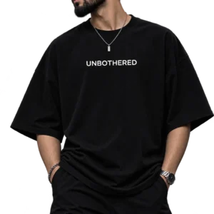 Male fashion model wearing an oversized black T-shirt with “UNBOTHERED” printed in white, styled with black cargo pants, sunglasses, a silver chain, watch, and ear piercing in a dark studio setting.