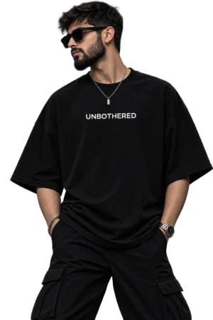Male fashion model wearing an oversized black T-shirt with “UNBOTHERED” printed in white, styled with black cargo pants, sunglasses, a silver chain, watch, and ear piercing in a dark studio setting.
